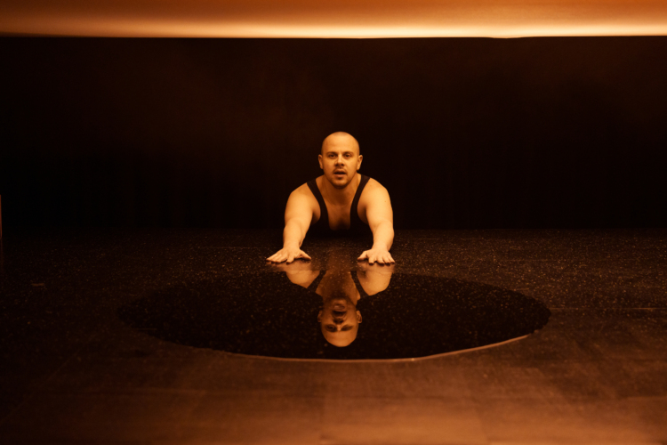 Un homme en débardeur noir se tient au sol, bras vers l'avant, au-dessus d'un miroir circulaire qui reflète son visage et le haut de son buste.