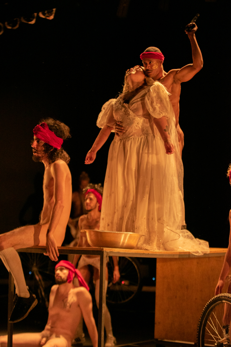 Sur une table, une personne en robe blanche se tient devant une personne nue avec un bandeau rouge sur la tête. D'autres sont assis·e·s dans l'espace avec un bandeau rouge sur la tête.