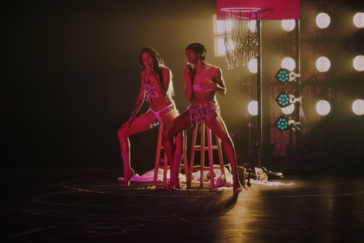 Assises sur des tabourets, et vêtues de bikini de corde blanche, Stacey et Satinée chantent au micro dans un espace au lumière tamisées.