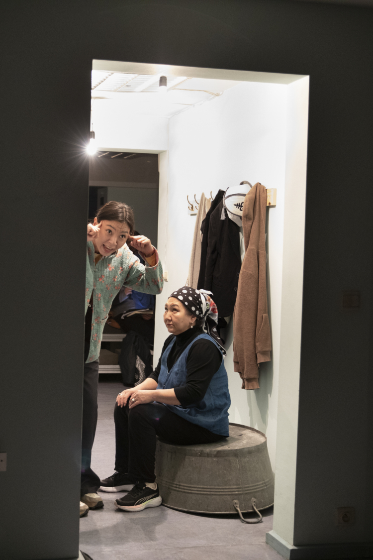 Une femme assise sur une bassine en métal regarde dans le vide alors qu'une autre femme s'adresse au public.