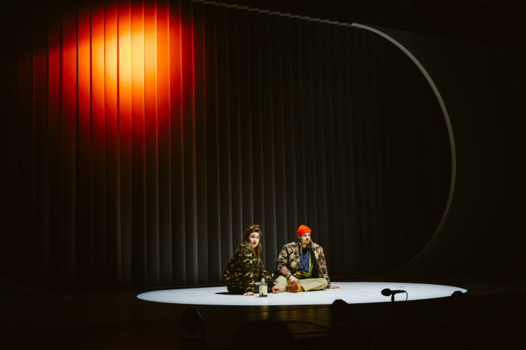 Querelle and Jezabel are sitting on the floor on stage, illuminated by a bright white light.