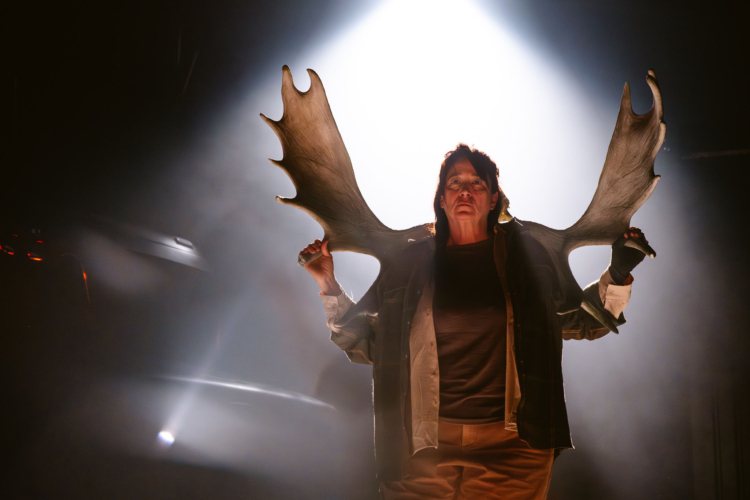 A male character holds large sticks behind his back which, with the effect of light, give the illusion that he has wings.