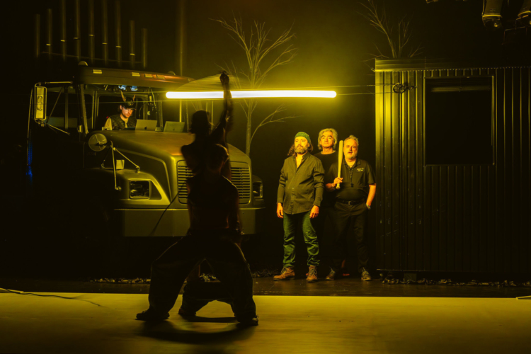 In front of a truck, on a stage lit in yellow, two silhouettes are naked against the light while three men in the background watch them.