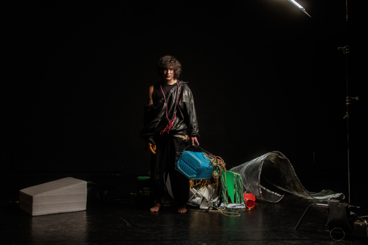 Un personnage vêtu de noir sur scène est au milieu d'un débarras d'objets et tient un panier qui contient un bidon bleu turquoise.