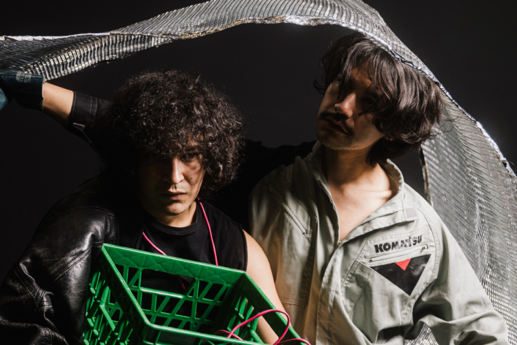 Un danseur portant une cagette verte, à gauche de l'image, et un autre en chemise de travail grise, à droite, se tiennent sous un tissu argenté.