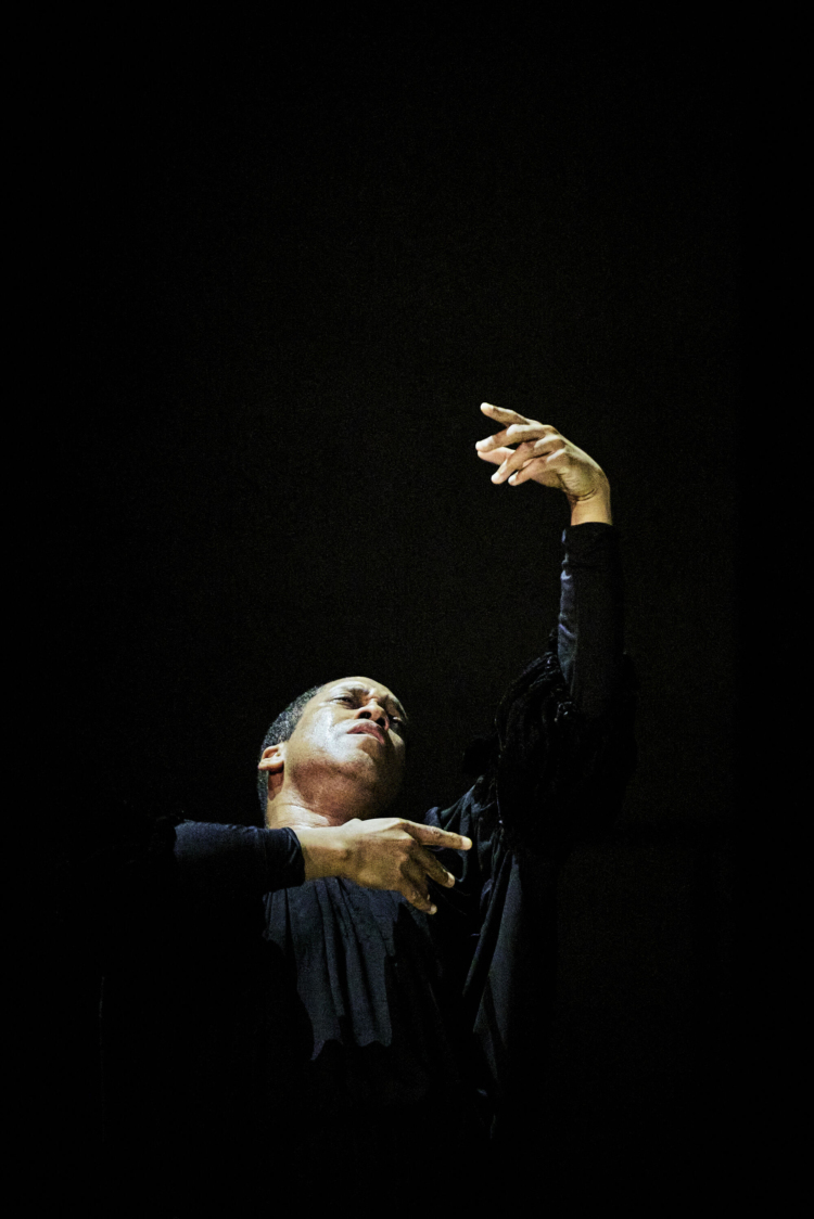 Choreographer Trajal Harrell, dressed in black, raises his left arm toward the sky and brings his right arm back toward his chest.