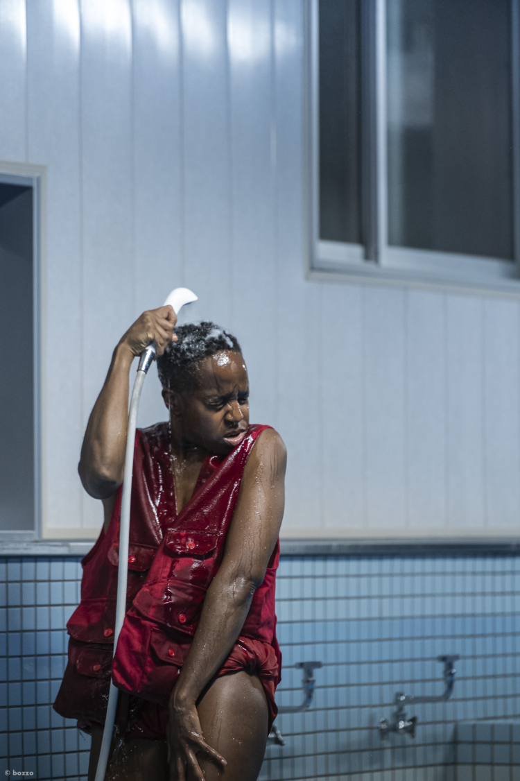 L'artiste Dana Michel vêtue d'un gilet rouge s'arrose la tête avec un pommeau de douche.