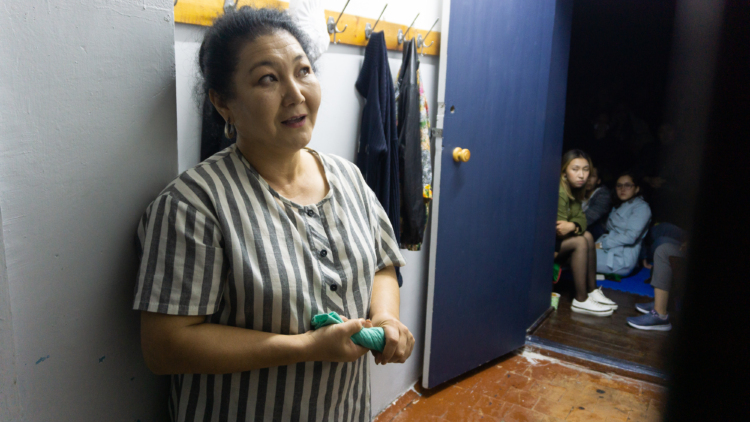 Une femme est accotée au mur. Elle parle, mais elle semble perdue dans ses souvenirs.