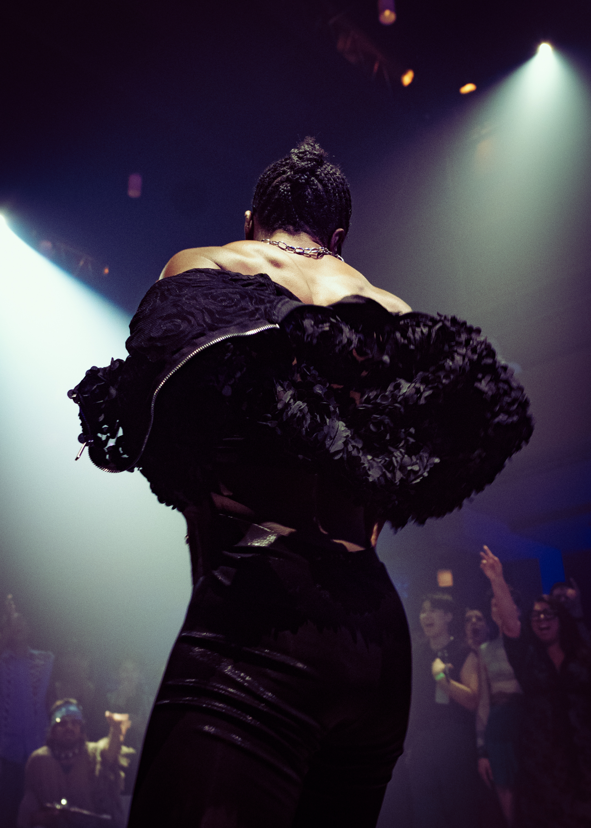 A person seen from behind, facing other people dancing under spotlights, is wearing a black puffy top.