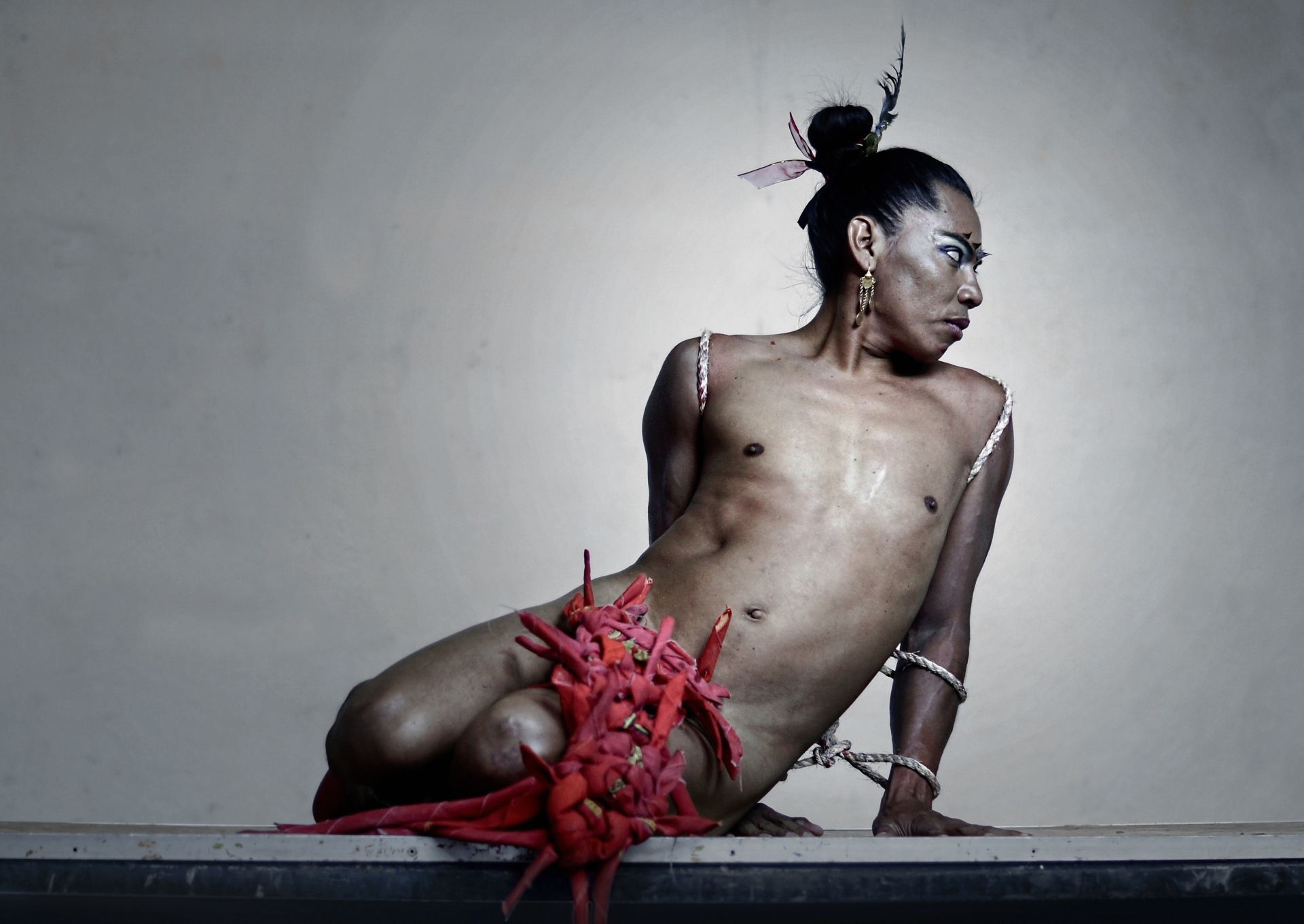 Lukas Avendano est assis sur sa cuisse gauche, s'appuyant sur sa main gauche, alors qu'il est presque nu, le sexe recouvert de morceaux de tissus rouges.
