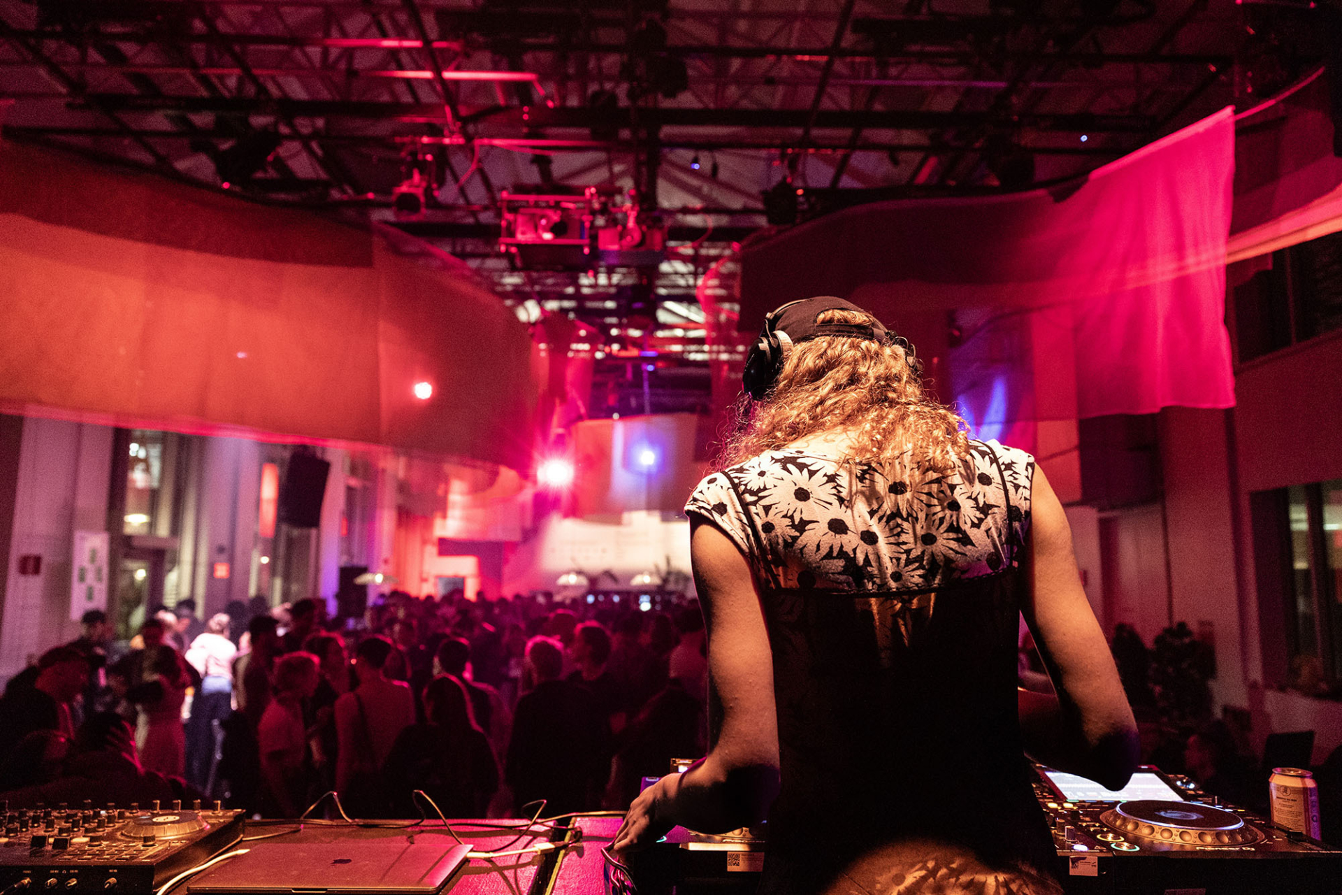 A person is DJing in front of a dancing crowd in a large room lit up in pink.