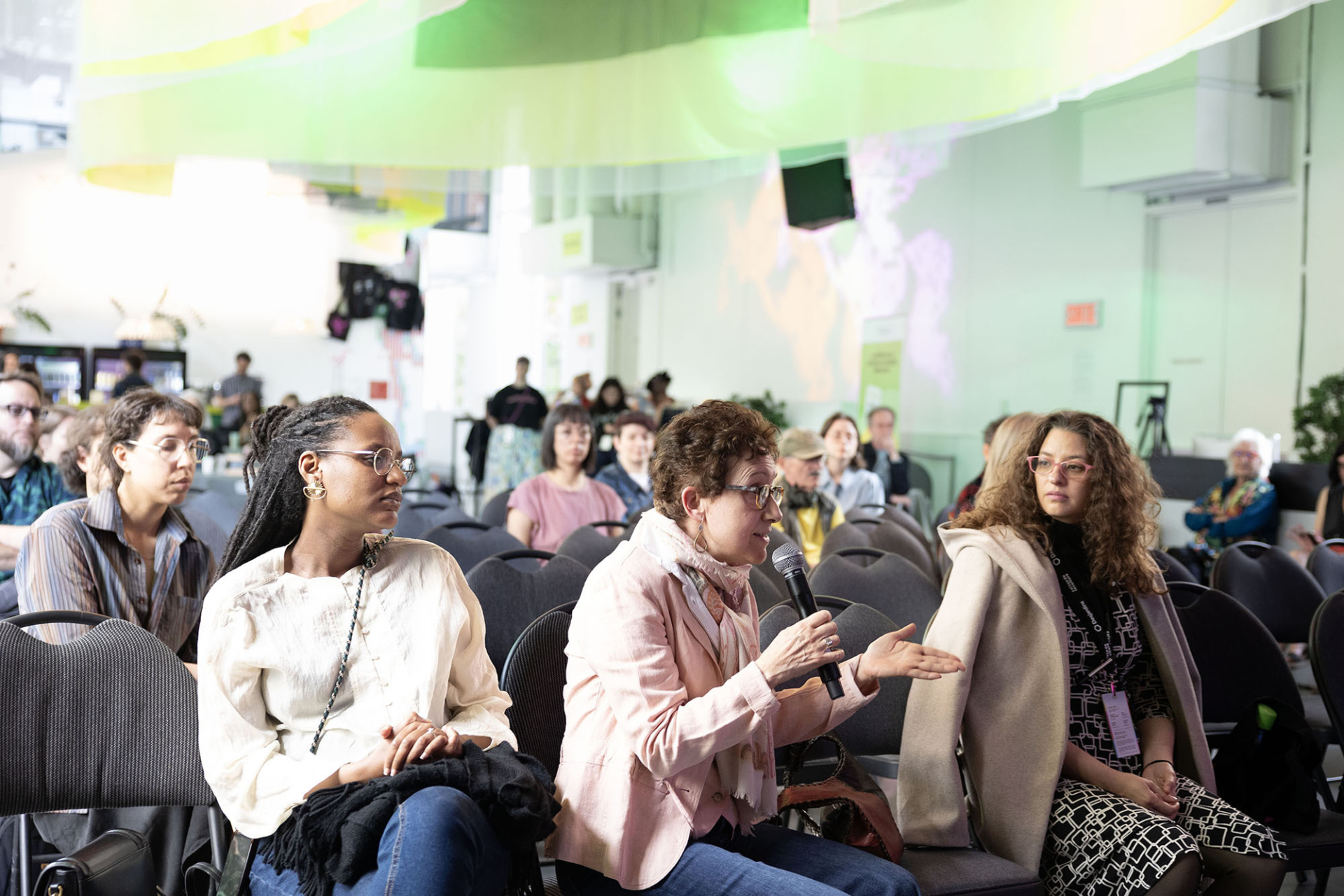 3 personnes sont assises au premier rang du public d'une conférence et celle du milieu prends la parole avec un micro.