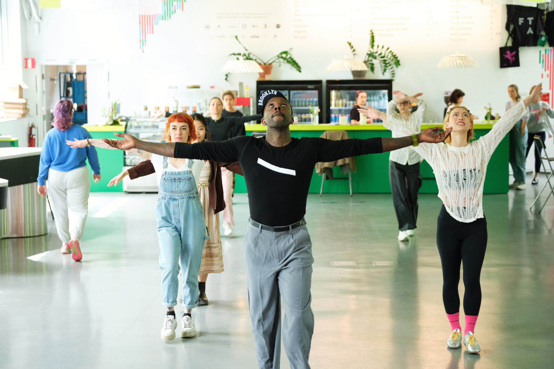 Dans une grande salle de réception, plusieurs personnes imitent le mouvement de Rja Feather Kelly qui avance les bras en croix.