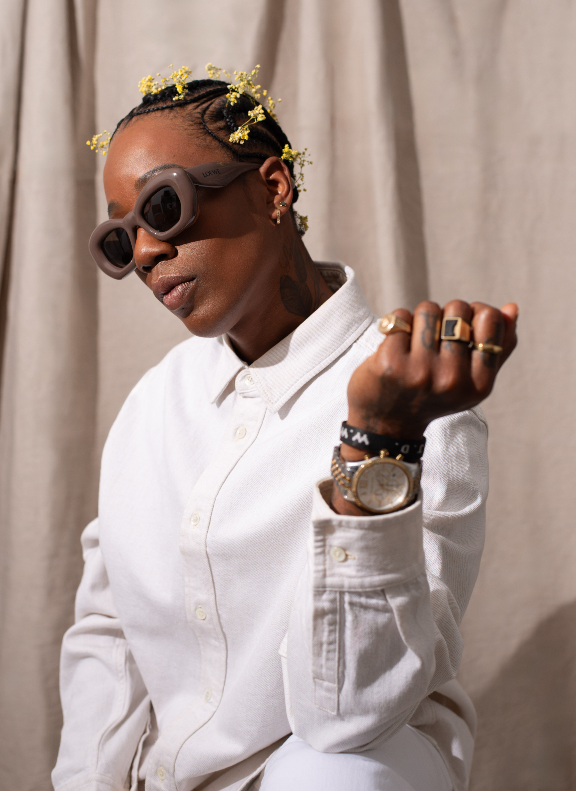 A portrait of A$h Banks wearing a white shirt and plum-colored sunglasses, posing in front of a beige curtain.