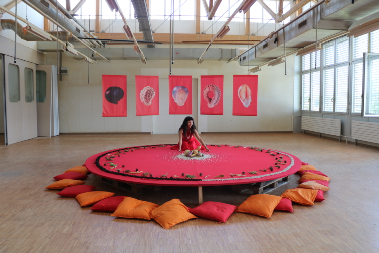 Taina Villalobos est assise sur un espace circulaire recouvert d'un tissu rose, légèrement surélevé et entouré de coussin au sol. Au centre et sur le pourtour du cercle, on aperçoit des semences et des graines.