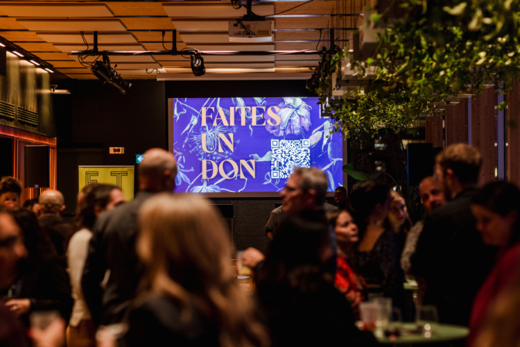 Plusieurs personnes sont réunis dans une salle pour un 6 à 8 de donateurs et donatrices et on aperçoit sur un écran l'image de la campagne de dons 2024-2025 du FTA avec écrit "Faites un don".