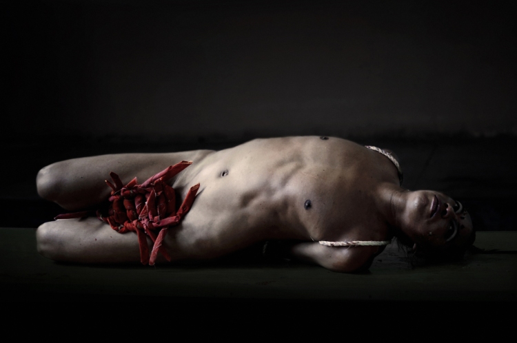 Lukas Avendano est allongé nu sur son flanc gauche alors que son entrejambe est recouvert de morceaux de tissu rouges.