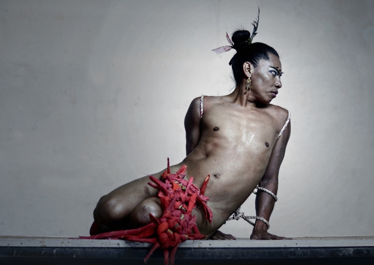 Lukas Avendano est assis sur sa cuisse gauche, s'appuyant sur sa main gauche, alors qu'il est presque nu, le sexe recouvert de morceaux de tissus rouges.