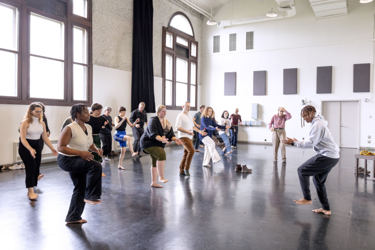 Des personnes du public sont dans une salle et suivent les mouvement montrés par Mamela Nyamza.