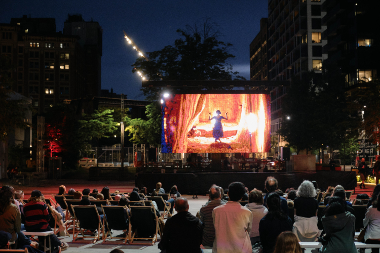 Un écran est installé en extérieur pendant la nuit et des spectateurs regardent le film qui y est projeté.