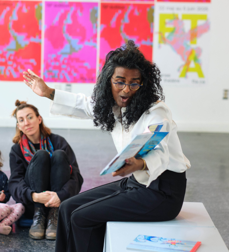 Kama La Mackerel tient un livre pour enfant dans les mains et en fait la lecture à des enfants.