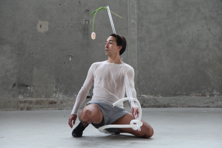 Stephen porte un t-shirt blanc et un short gris dans un espace en béton et tient dans sa main gauche une grande tige en plastique transparent avec une tulipe d'un bord, et un tuyau blanc noué de l'autre.