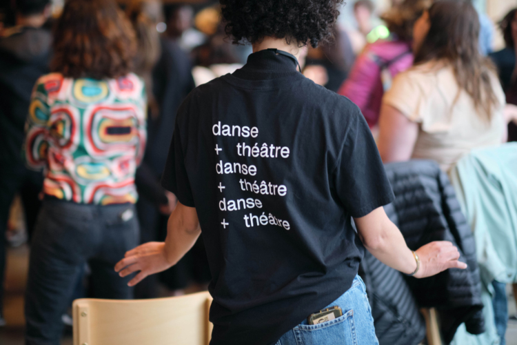 Une personne de dos porte un t-shirt noir où il est inscrit trois fois "danse + théâtre".